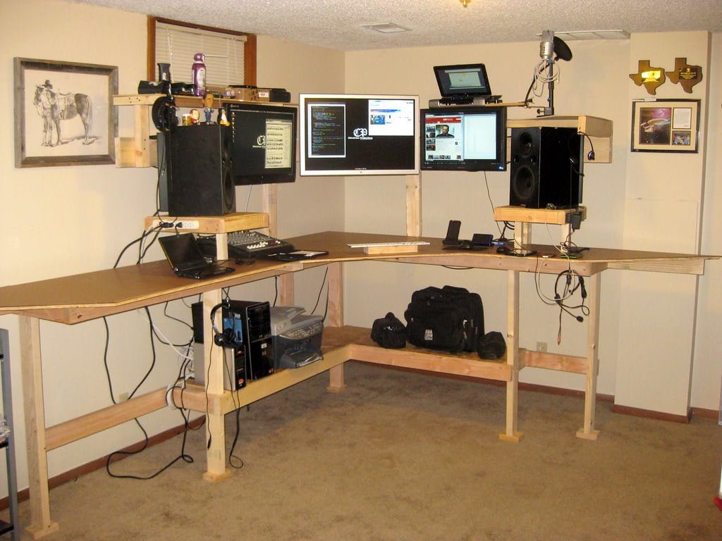 A DIY home office setup featuring a wooden corner desk with multiple monitors raised on makeshift stands, speakers, a laptop, and various electronic equipment, with cables visible and personal items scattered about.