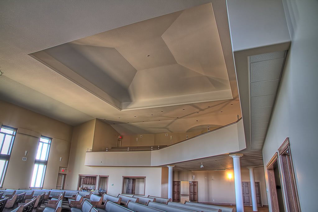 Interior view of a modern hall with high ceilings, rows of benches, a balcony level, large windows, and a complex geometric ceiling design.