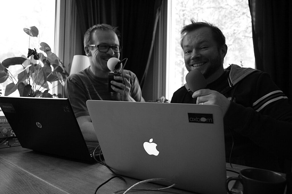 Two men laughing and holding microphones while sitting at a table with laptops in a room with a plant and window in the background, black and white photo.