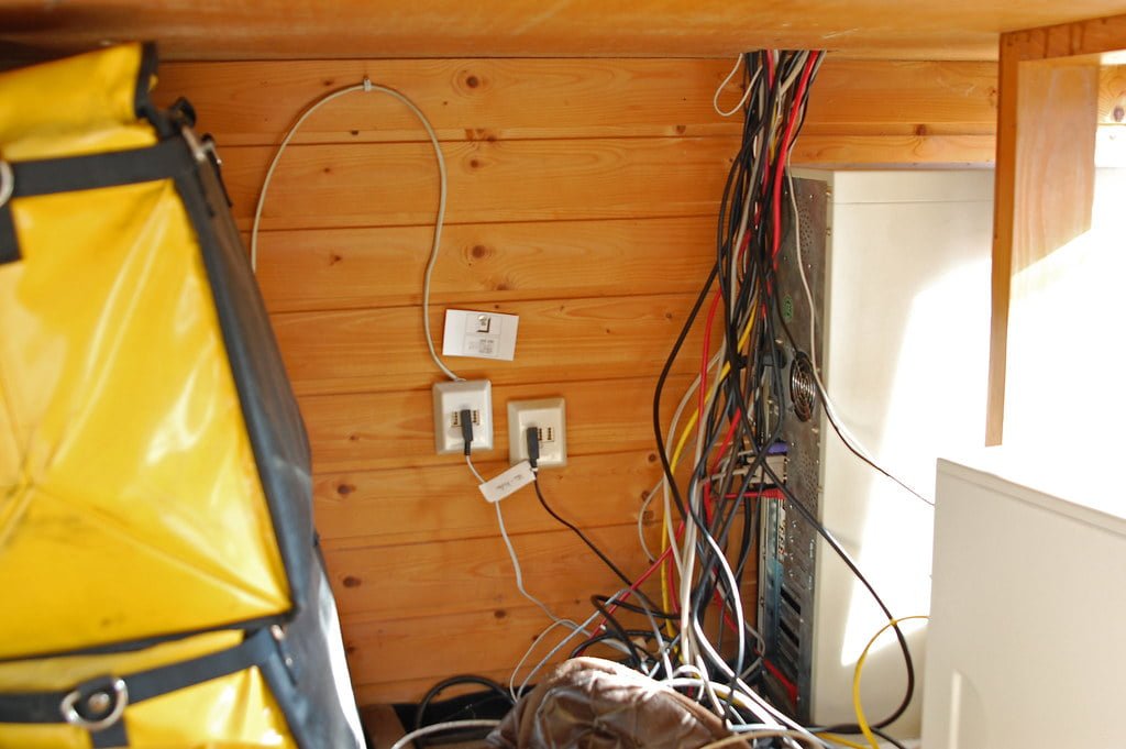 An image showing a cluttered corner with exposed wires and cables near an electrical panel, against wooden paneling, with a bright window to the side and a yellow bag in the foreground.