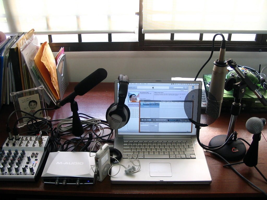 A home recording setup on a desk with a laptop running audio editing software, an audio interface, a mixer, two microphones with stands, headphones, and some file folders to the side.