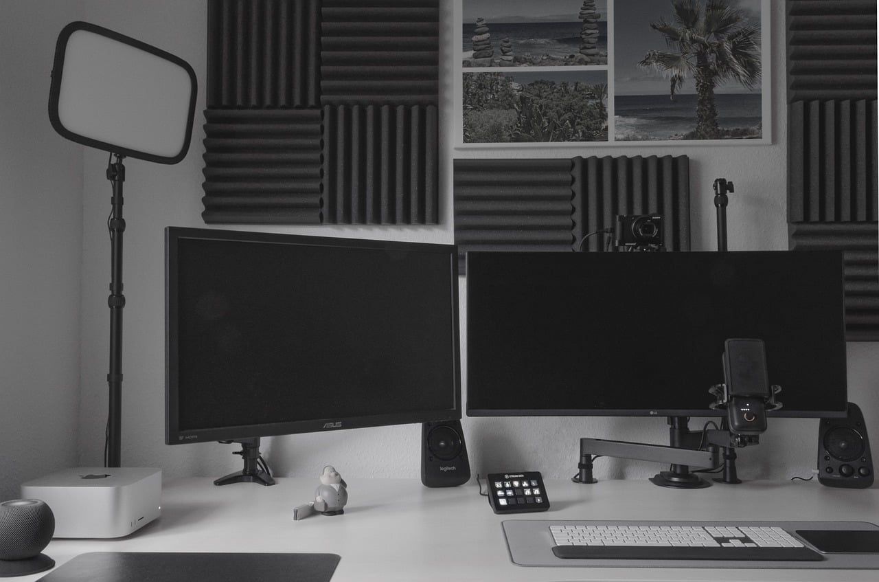 A grayscale image of a modern workspace featuring two off computer monitors, a keyboard, a mouse, a pair of speakers, a smartphone on a stand, and a small figurine. In the background, there's acoustic foam on the wall with framed pictures above and a camera on a tripod to the side. A LED light panel on a stand is visible to the left. The setting suggests a neat, professional home office environment.