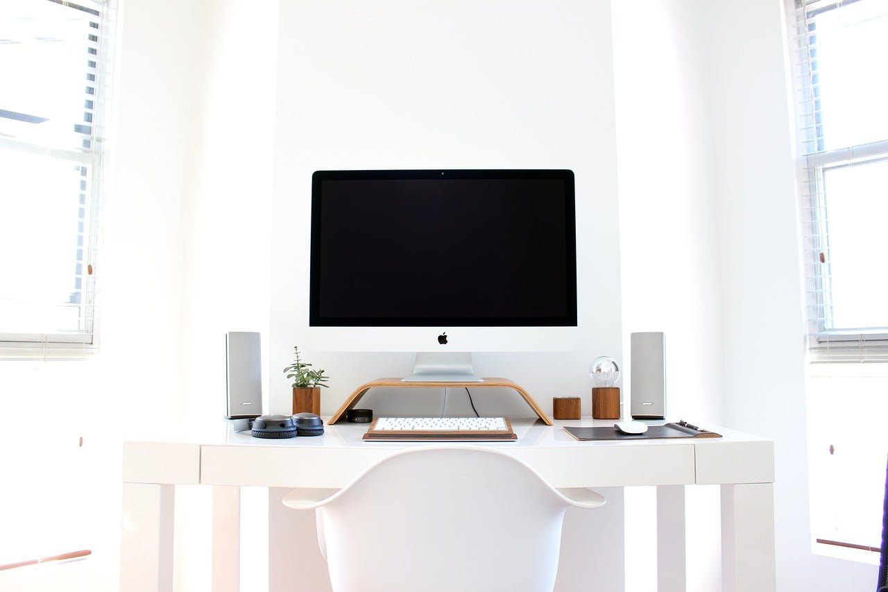 A bright and minimalist home office setup with an iMac on a white desk, flanked by speakers and accessorized with a small potted plant, a wooden stand, and headphones.