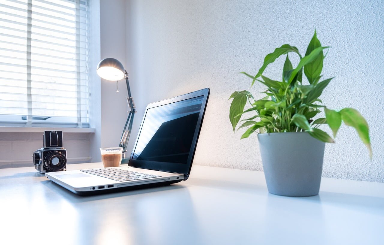 A laptop on a desk beside a vintage camera and a glass of iced coffee with a potted plant on the right and a desk lamp turned on in the background near a window with blinds.