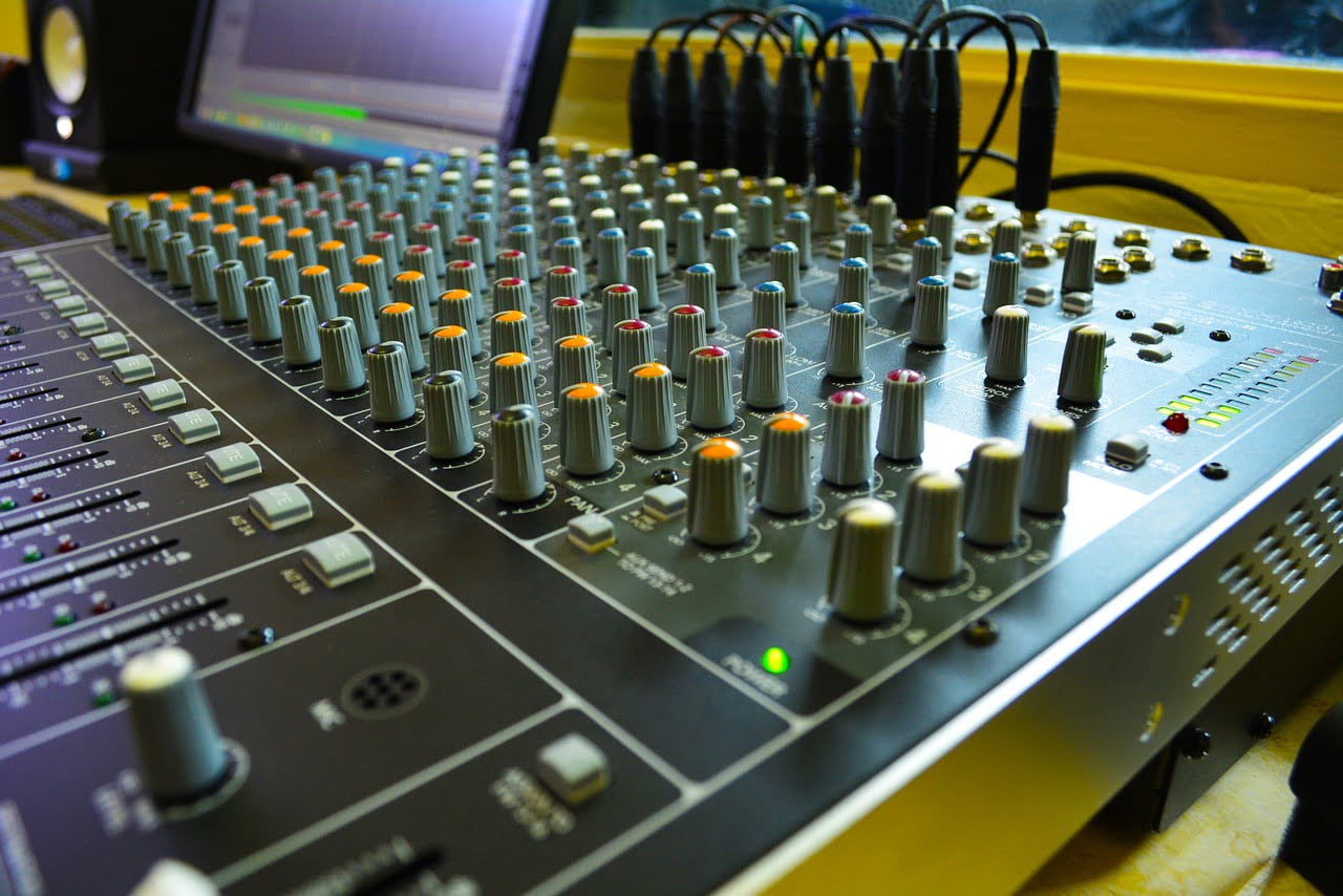 Close-up of an audio mixing console with various knobs and sliders, some with colored indicators, and cables connected at the back, in a studio environment.