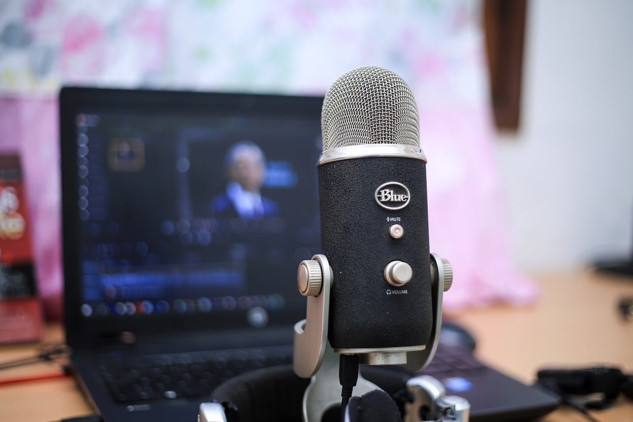 A Blue Yeti microphone in focus on a desk with an out-of-focus laptop displaying a video editing application in the background.