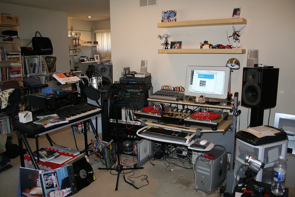 A home music studio with electronic equipment including keyboards, mixers, monitors, turntables, and a computer, amidst a cluttered room with books, figurines on shelves, and CDs strewn about.