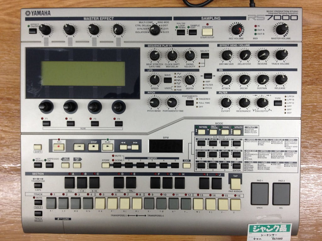 Yamaha RS7000 Music Production Studio equipment featuring knobs, buttons, and sliders for various functions such as sampling, master effects, and sequencing, with a large LCD screen in the center, on a wooden surface.