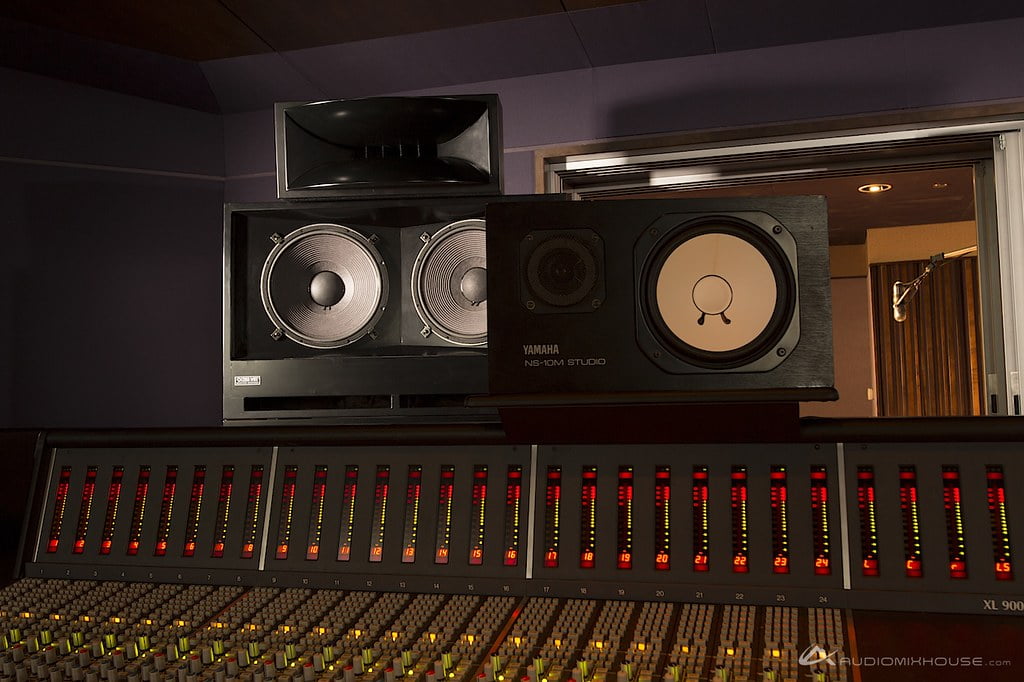 A professional recording studio control room with a large mixing console in the foreground with lit channel indicators, and Yamaha NS-10M studio monitors stacked atop larger speakers in the background.