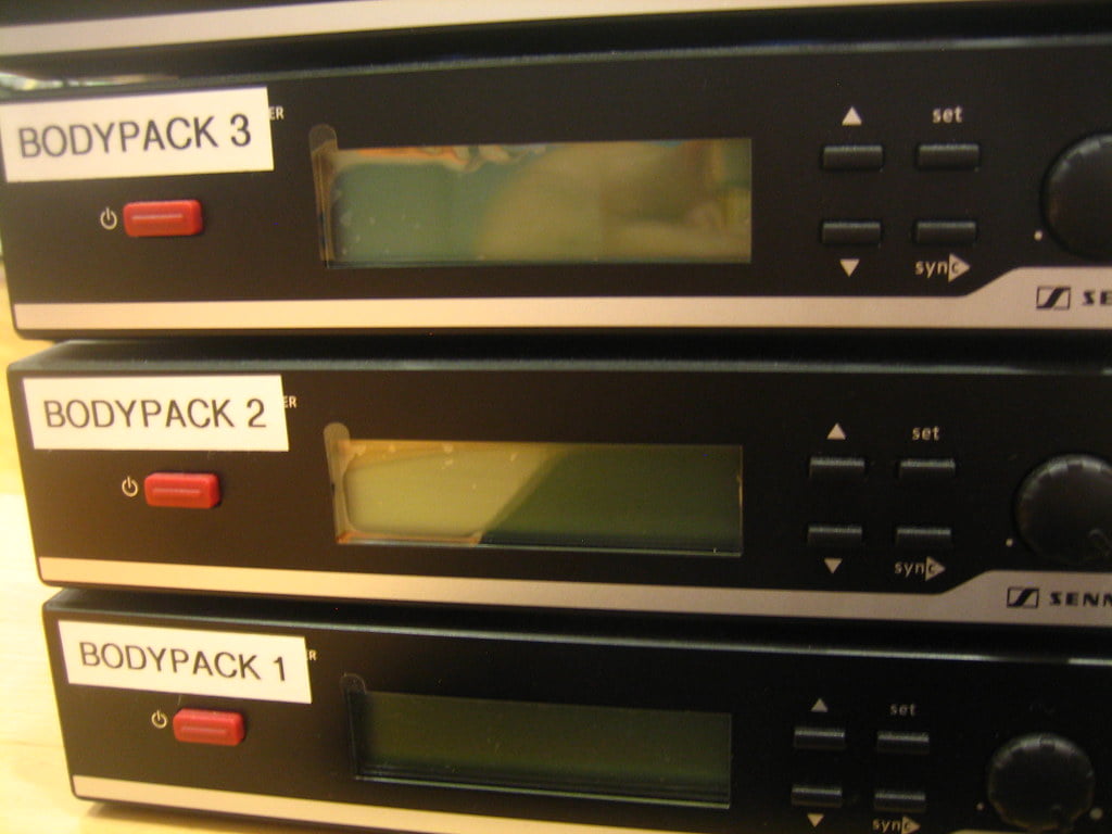 A close-up of three audio bodypack transmitters stacked on top of each other, labeled BODYPACK 1, BODYPACK 2, and BODYPACK 3, with LED power indicators and sync buttons, and the brand "Sennheiser" visible on the bottom device.