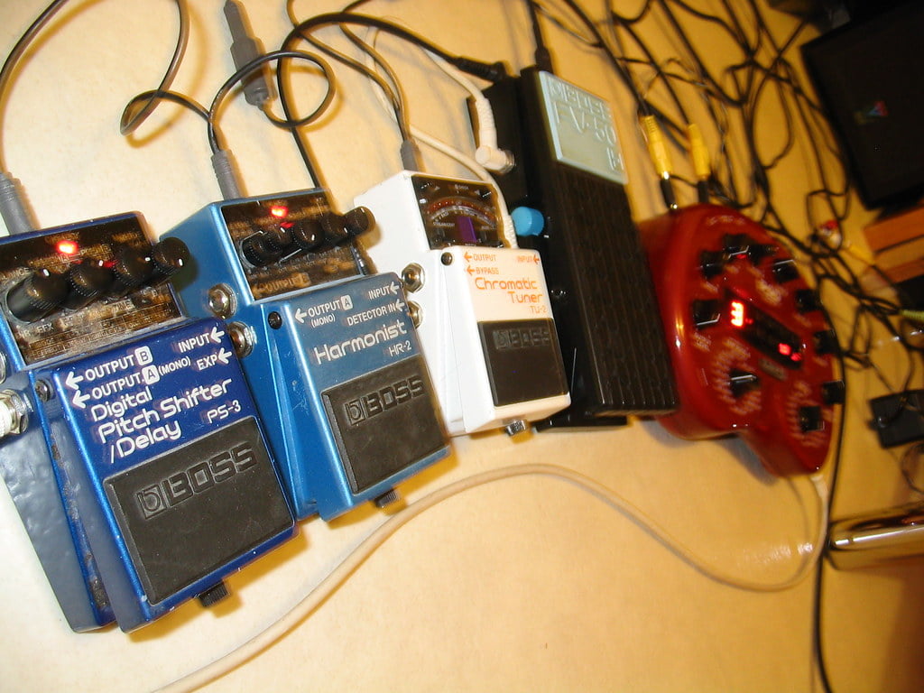 A collection of guitar pedals including a digital pitch shifter/delay, a harmonist, and a chromatic tuner connected by cables on a floor, with a blurred background hinting at a musical setting.