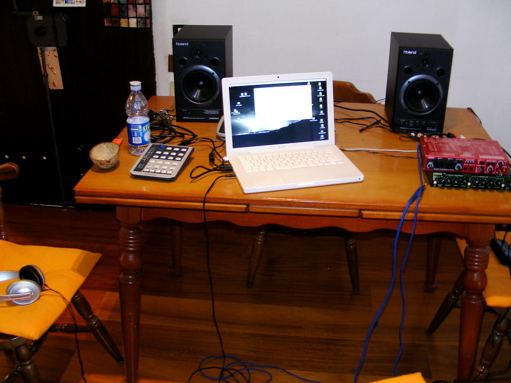 A laptop set up on a wooden table flanked by two studio monitors with various audio equipment and cables, a calculator, and a water bottle also on the table.