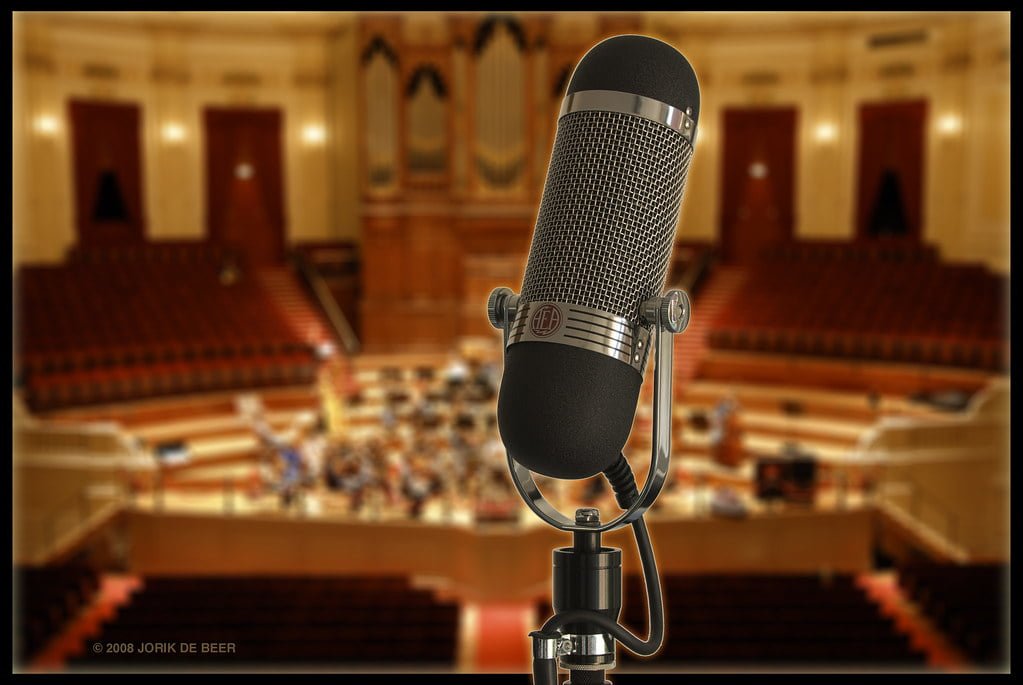 A vintage microphone in sharp focus set against a blurred background of an empty concert hall with red seats and elegant decor.