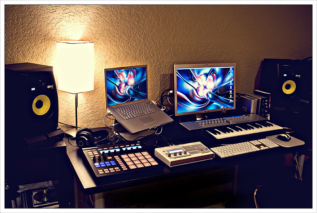 A home music production setup with two monitors displaying colorful abstract wallpapers, studio monitor speakers, a MIDI keyboard, pad controller, laptop, and a lamp on a desk.