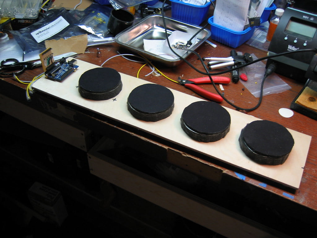 A wooden board with four black hockey pucks on a cluttered workbench, surrounded by various tools and electronic components.