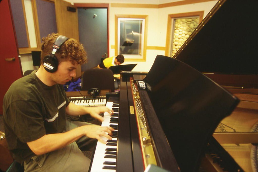 A man wearing headphones playing a grand piano in a recording studio, with another person visible in the background through the studio window.