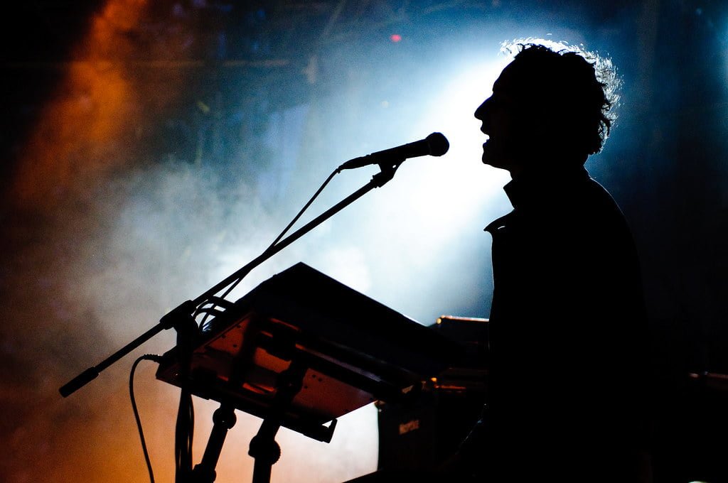 Silhouette of a musician singing into a microphone and playing a keyboard on a stage with dramatic backlighting and haze.