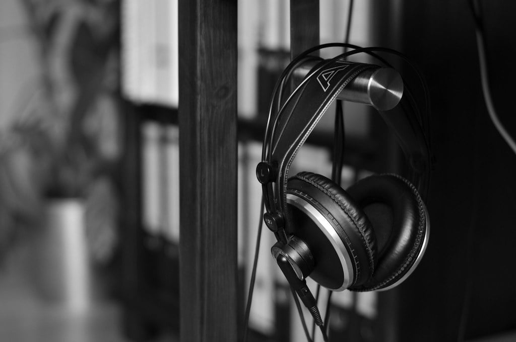 A pair of black over-ear headphones hanging on a metal hook attached to a wooden bookshelf, with blurred books in the background, in a black and white image.