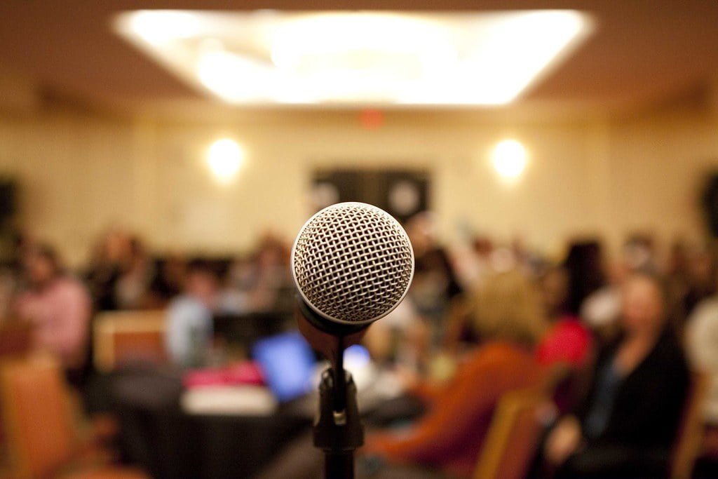 Close-up of a microphone in focus with a blurred audience in the background in a conference room.