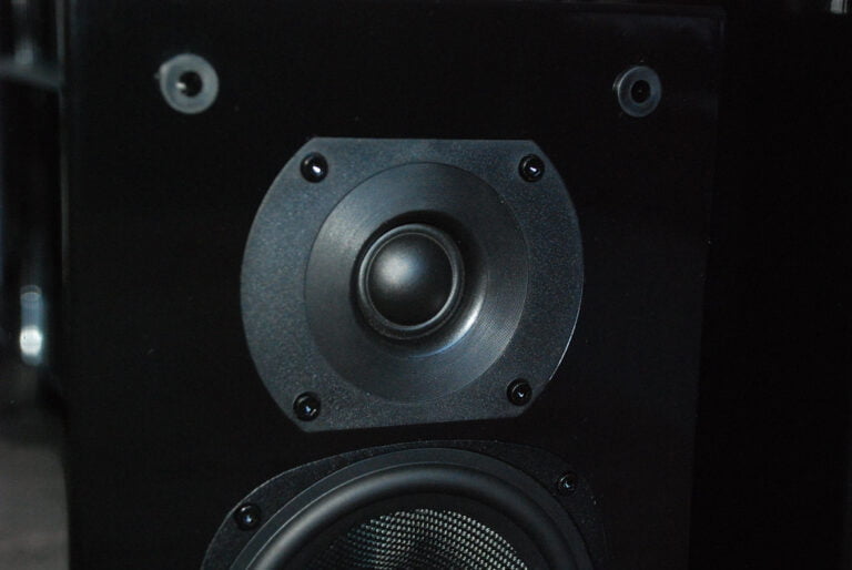 A close-up of a black speaker cabinet showing the tweeter at the top, the mid-range speaker in the center, and the top edge of the woofer at the bottom, all mounted on a matte surface with visible screw heads.