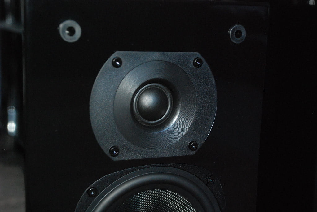 A close-up of a black speaker cabinet showing the tweeter at the top, the mid-range speaker in the center, and the top edge of the woofer at the bottom, all mounted on a matte surface with visible screw heads.