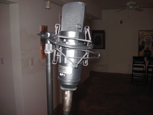 A condenser microphone with a shock mount attached to a metal stand in a room with artwork on the wall in the background.