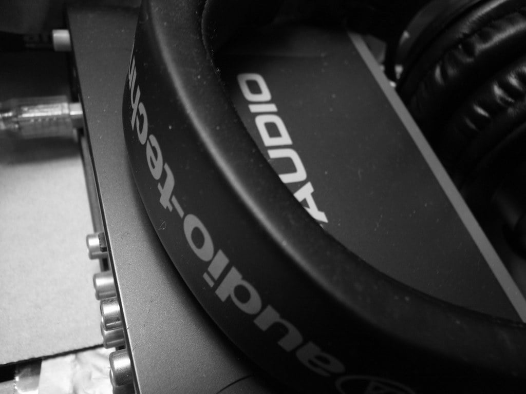 Black and white close-up photo of headphones with the brand name "AUDIO" visible on its band, resting on a surface alongside audio equipment.