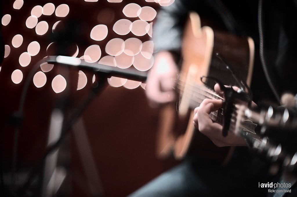 A close-up of a person playing an electric guitar with a blurred background of bokeh lights, with a microphone stand slightly in view.