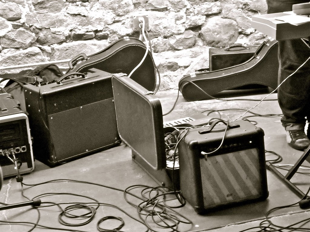 A black and white photo of a cluttered music stage with various guitar amplifiers, cables, and instrument cases.