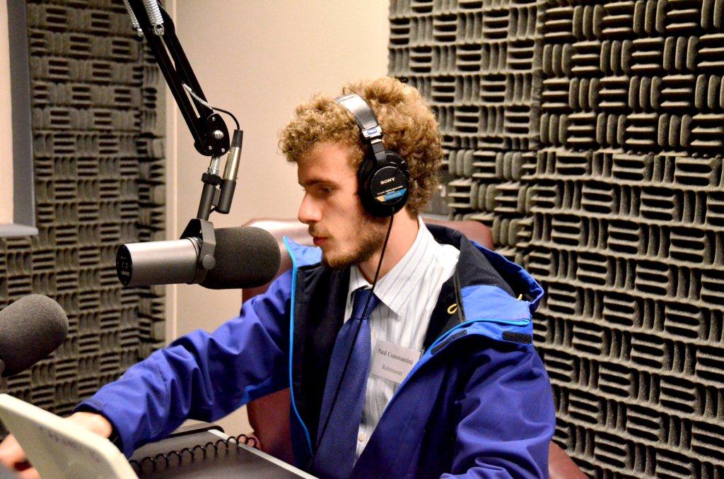 A man with curly hair wearing headphones speaks into a microphone in a soundproofed radio studio while reading from a document.