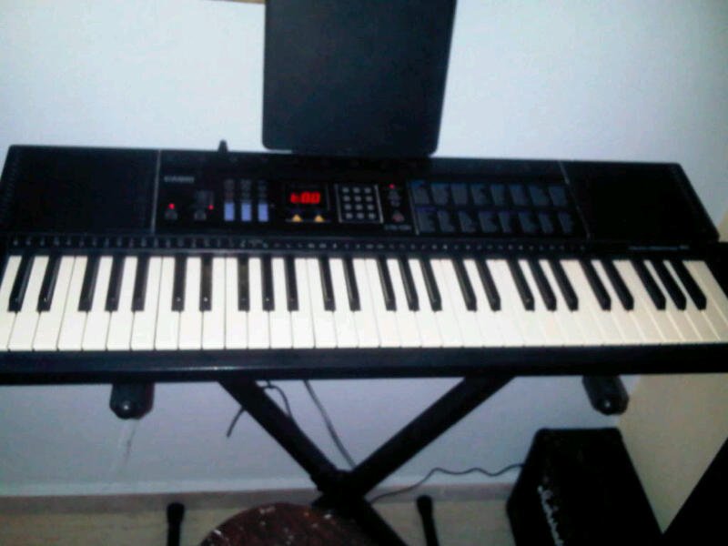 An electronic keyboard on a stand with a speaker to the right, against a white wall in a dimly lit room.