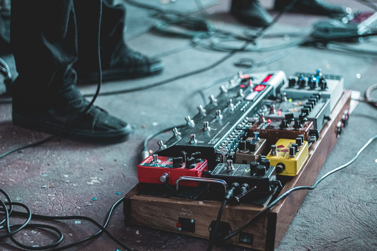 A collection of guitar effect pedals on a pedalboard with cables, set on a stage floor with a musician's legs and shoes visible in the background.