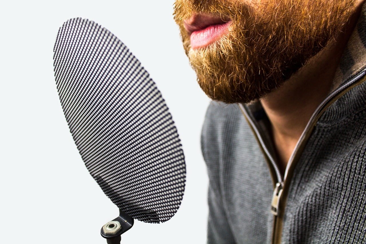 Close-up of a bearded man speaking into a microphone with a pop filter, isolated on a white background.