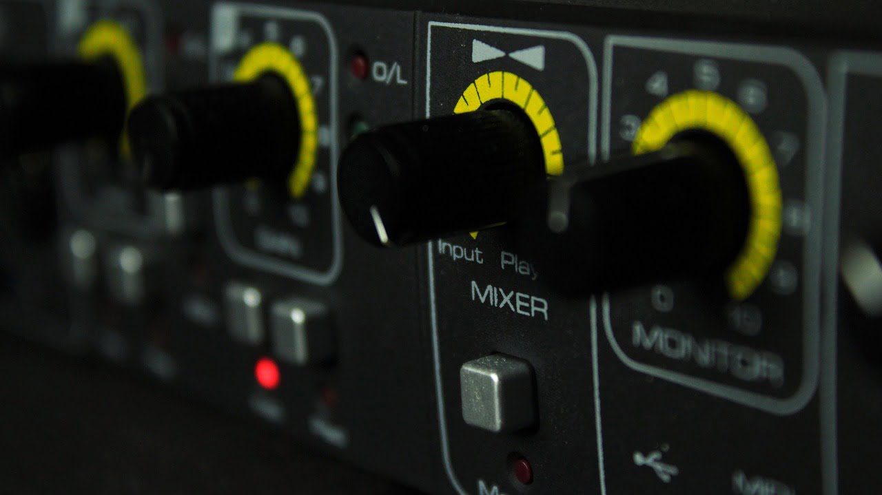 Close-up of an audio mixer's control panel with knobs and buttons, highlighting the input level and monitor controls with yellow accentuated levels, shot in low light.