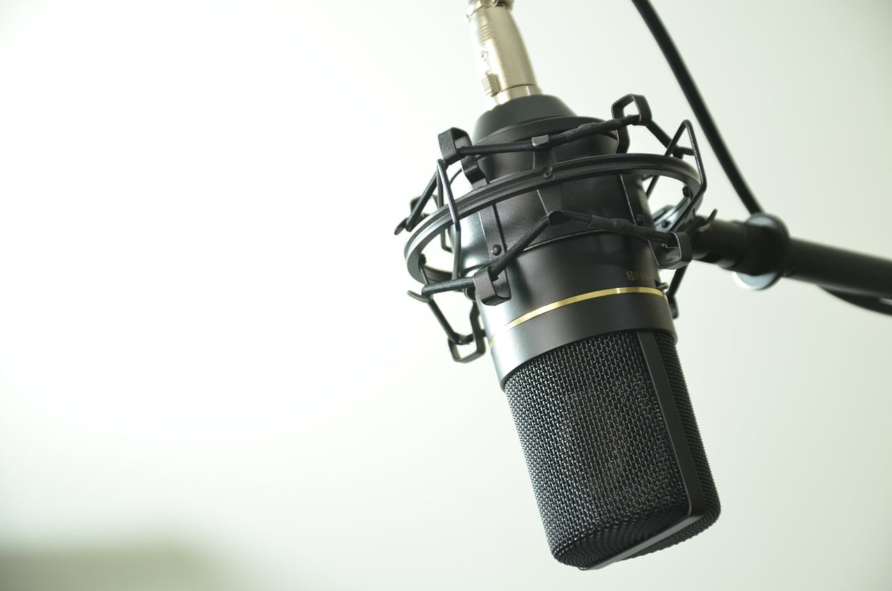 A close-up of a studio microphone suspended on a boom arm against a blurred light background.