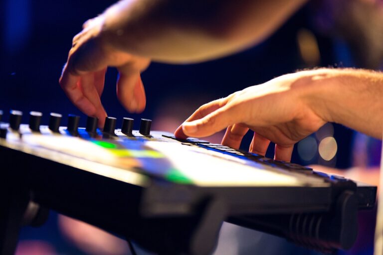 Hands playing a digital keyboard with illuminated buttons and sliders on a blurred stage background.