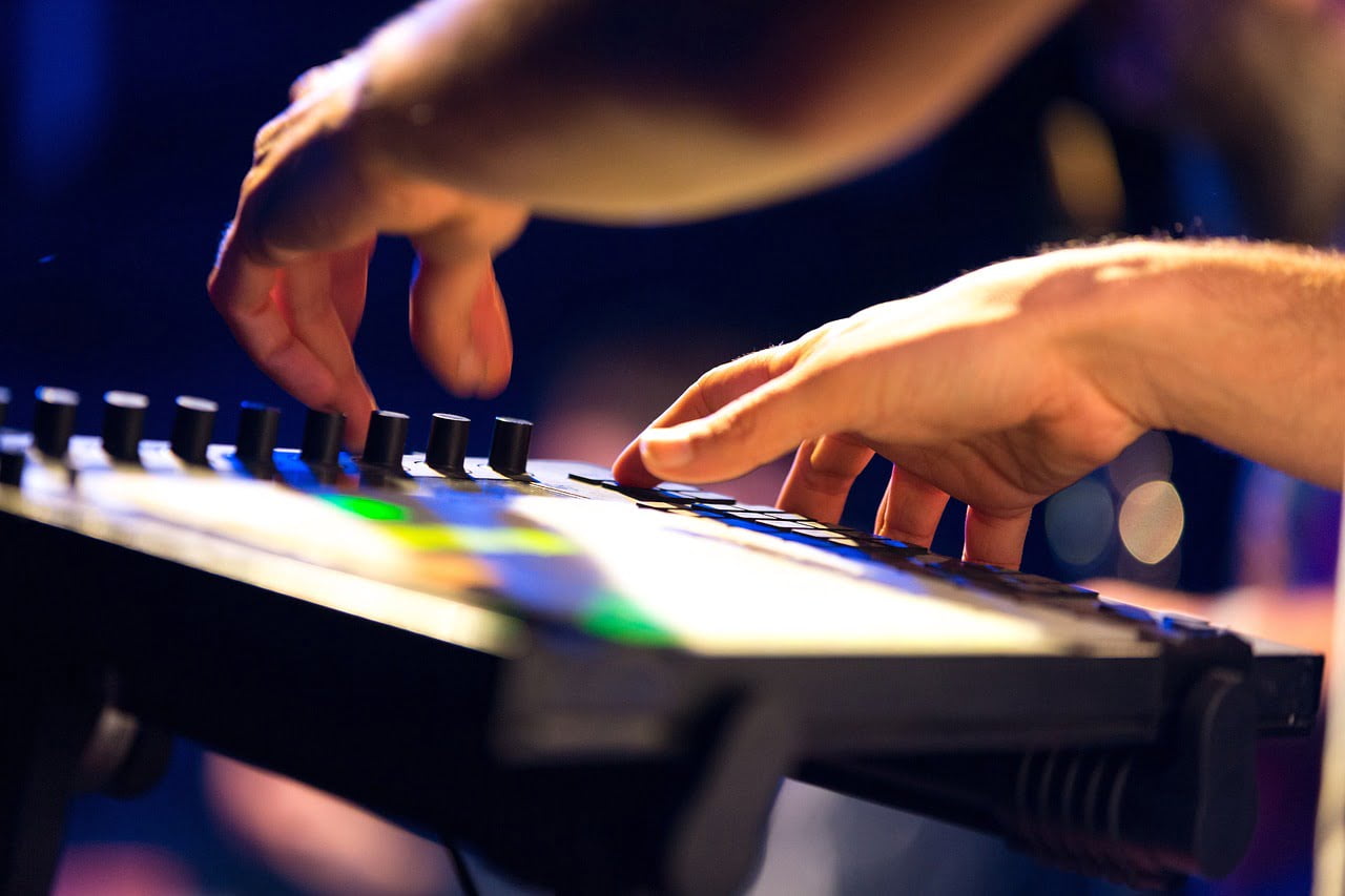 Hands playing a digital keyboard with illuminated buttons and sliders on a blurred stage background.