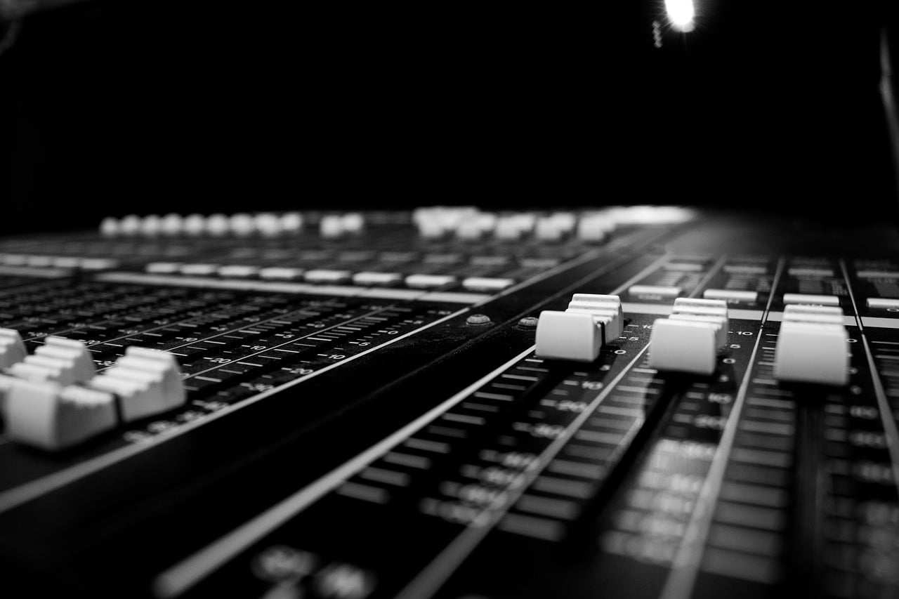 A black and white close-up photo of an audio mixing console with multiple sliders and knobs, focused on the central part of the board.