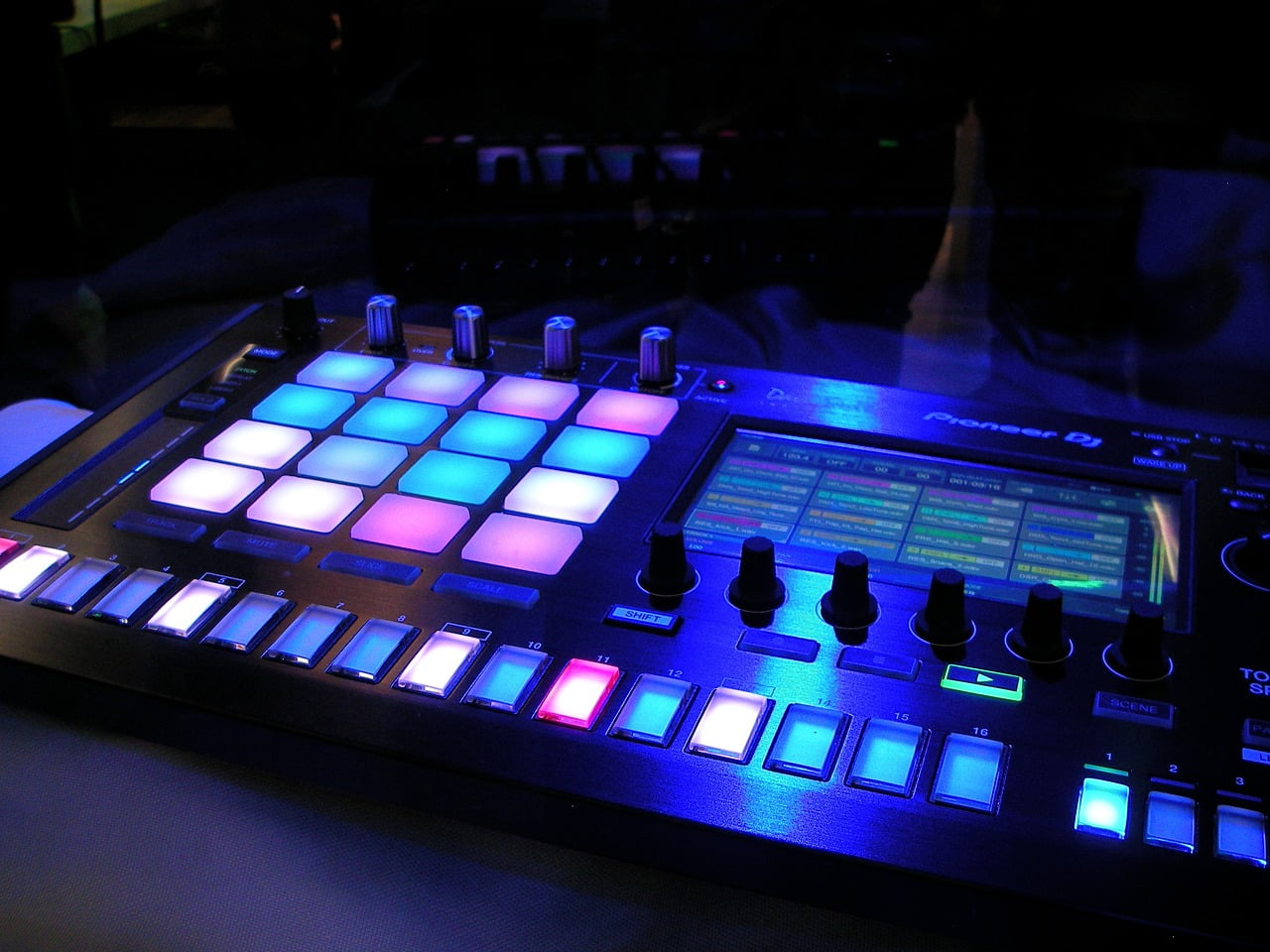 Illuminated DJ controller with pads, knobs, and sliders, displaying various colors in a dark setting.